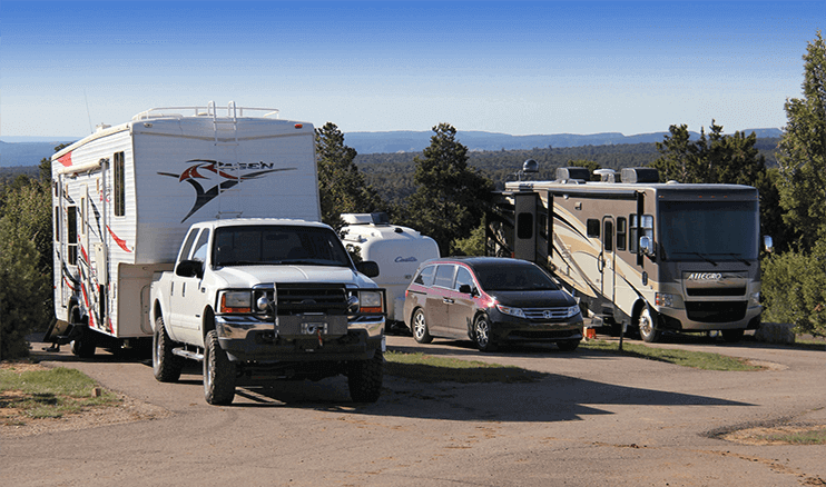 A white RV, a truck, and a silver car are parked at a scenic campsite surrounded by trees.